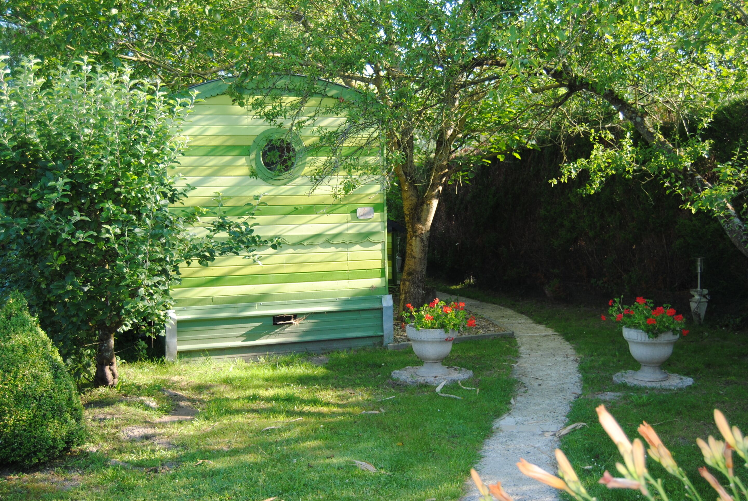 Una estructura de madera verde con un ventanal circular, rodeada de abundante vegetación y un sendero de grava.