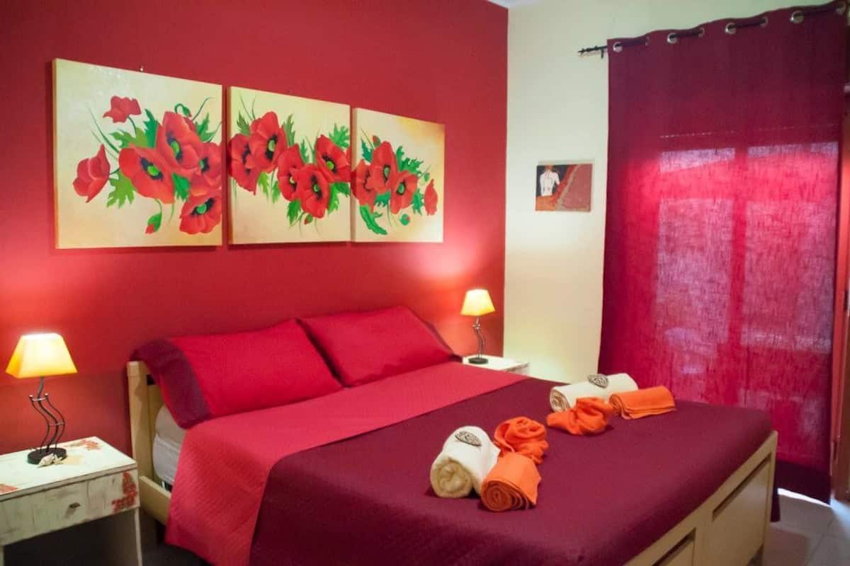 A hotel room with a red bedspread, two wall-mounted paintings of red flowers, a red curtain, and a nightstand with a lamp and a folded towel.