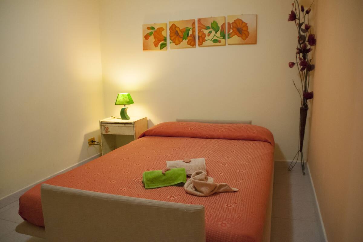A neatly made bed with an orange bedspread, a green towel, and a beige towel on the bed. There is a nightstand with a lamp and a vase with flowers beside the bed.