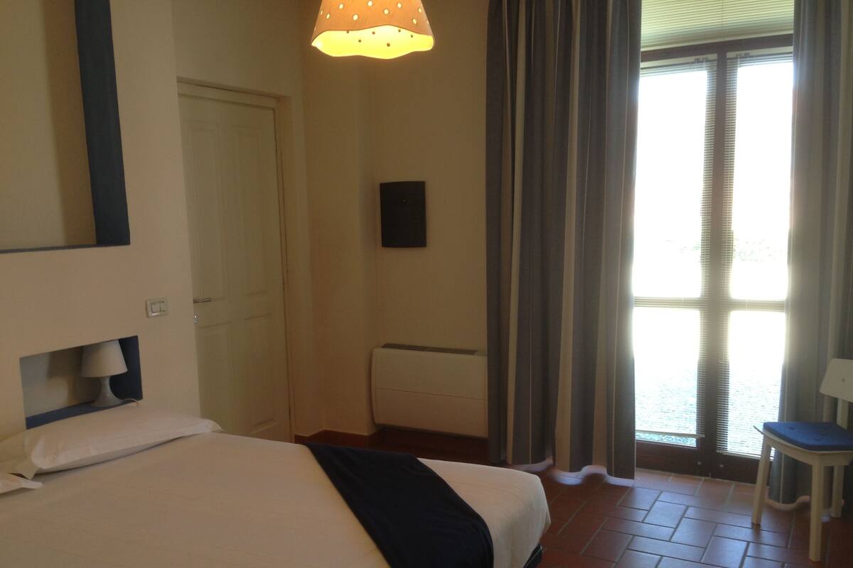 A hotel room with a bed, a bedside table, a lamp, a window with curtains, and a view of the sea.