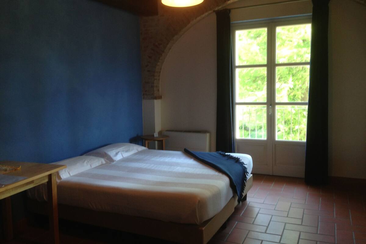 A bedroom with a bed, a table, and a window with a view of greenery.