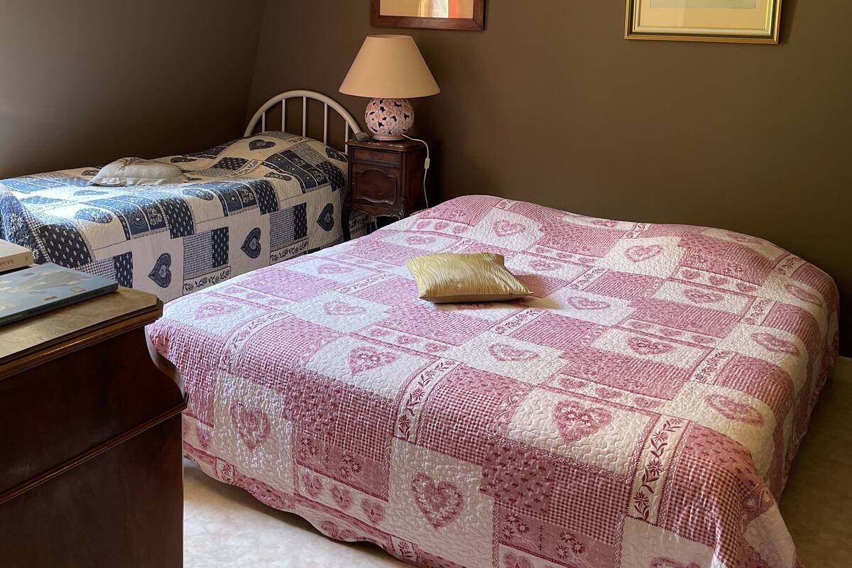 Ein Schlafzimmer mit zwei Betten, eines mit einem gemusterten Bettbezug und das andere in einfarbigem Design. Ein Nachttisch mit Lampe, ein gerahmtes Bild an der Wand und ein Fenster, das für natürliches Licht sorgt.