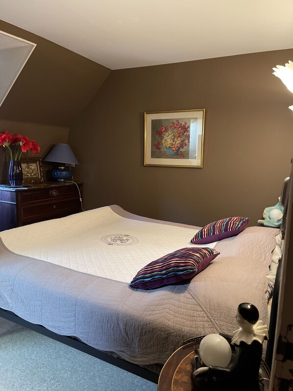 Een slaapkamer met een bed, kussens, een dressoir, een lamp, een ingelijste foto en een vaas met bloemen.