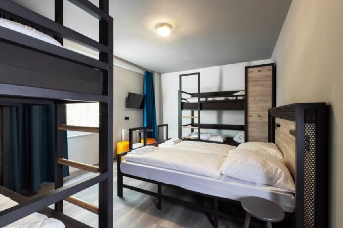 Een slaapkamer met stapelbed, televisie, een klein krukje en een raam met gordijnen.