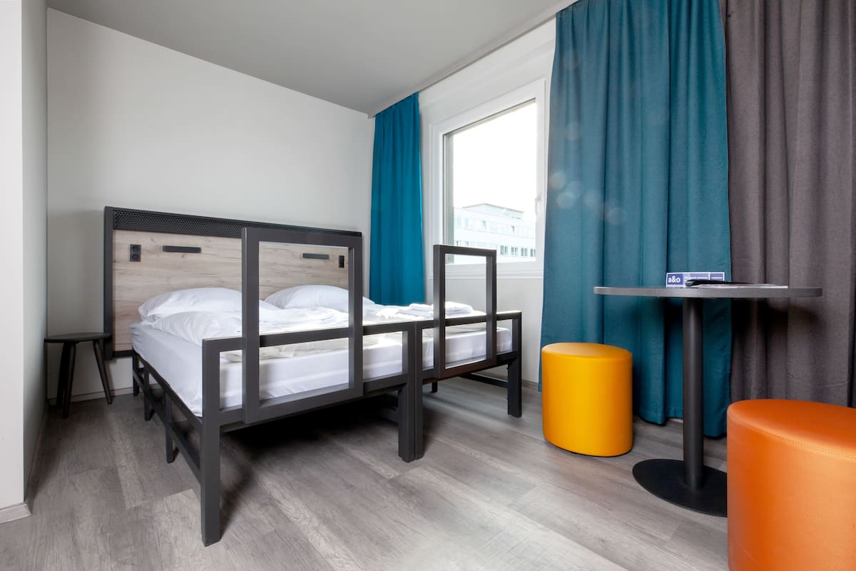 Une chambre d’hôtel moderne avec un lit en bois, une petite table, un pouf jaune et une fenêtre avec des rideaux.