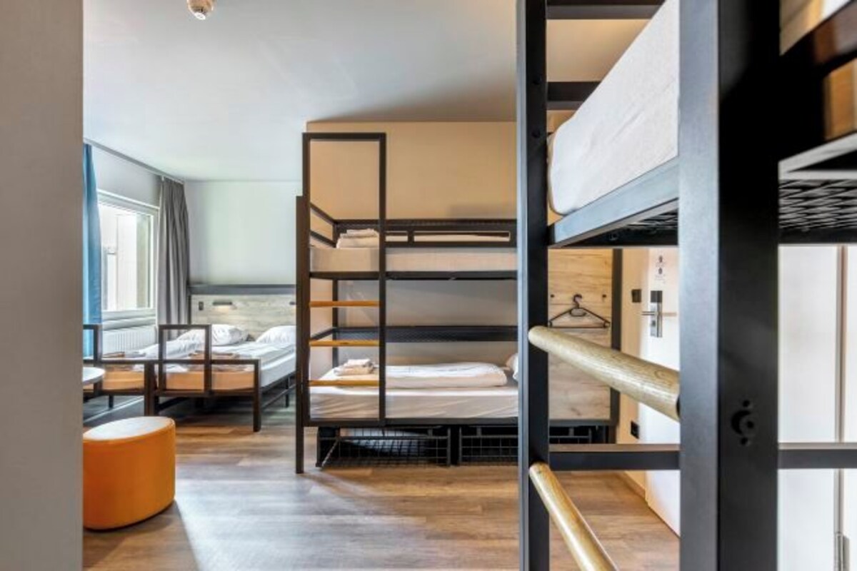 Een slaapkamer met stapelbedden, een houten vloer, een klein oranje poef en een raam met gordijnen.