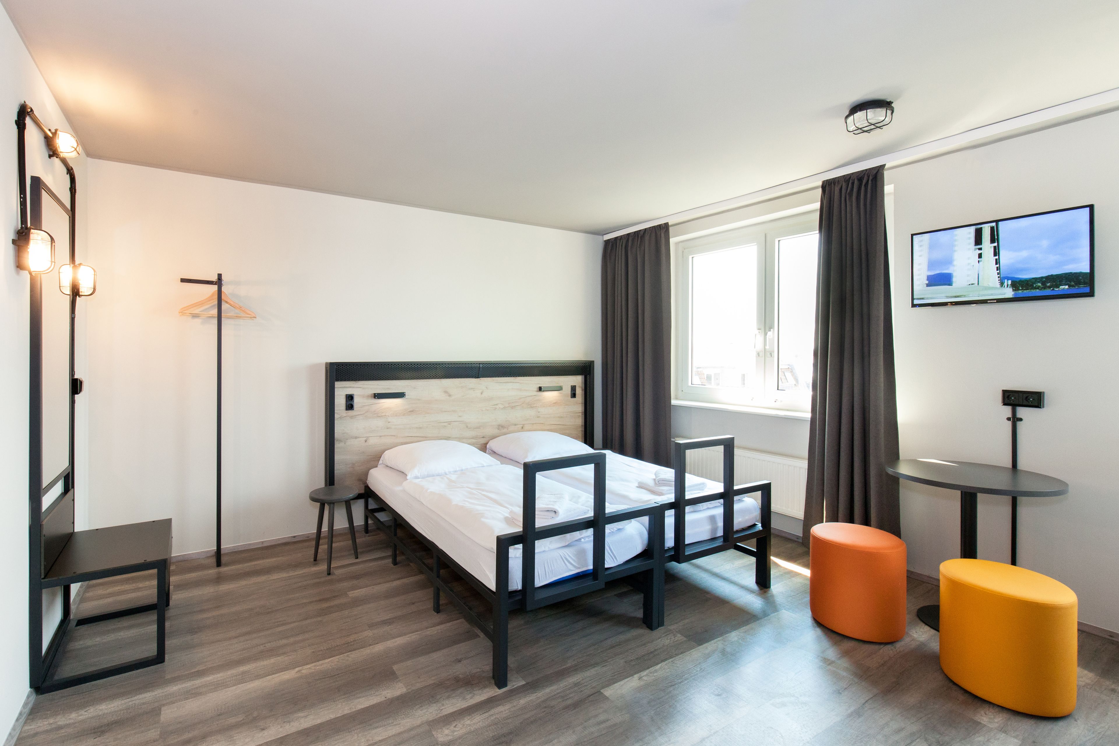 Ein modernes Hotelzimmer mit einem Bett mit Holz Kopfteil, einem kleinen Tisch, zwei Hocker, einem an der Wand montierten Fernseher und einem Fenster mit Vorhängen.
