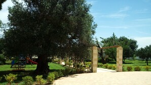 A paved entrance area with a large tree and a playground in the background. - Residence Gli Ulivi (Leverano)