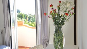 A room with a white table, a clear vase with flowers, a white chair, and a view of a garden through a glass door. - Bellatrix (Portici)