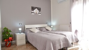 A bedroom with two beds, a nightstand with a lamp, a potted plant, and a wall-mounted air conditioner. - Bellatrix (Portici)