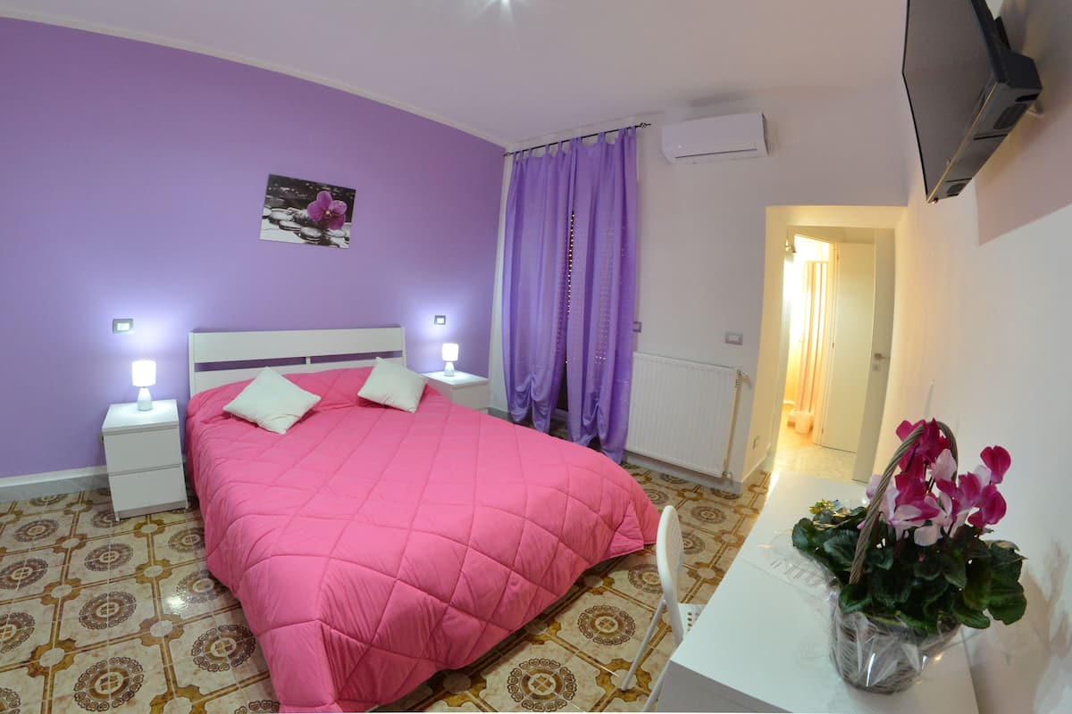 A bedroom with a pink bedspread, white nightstands, a bedside lamp, a purple curtain, and a potted plant on a white table.