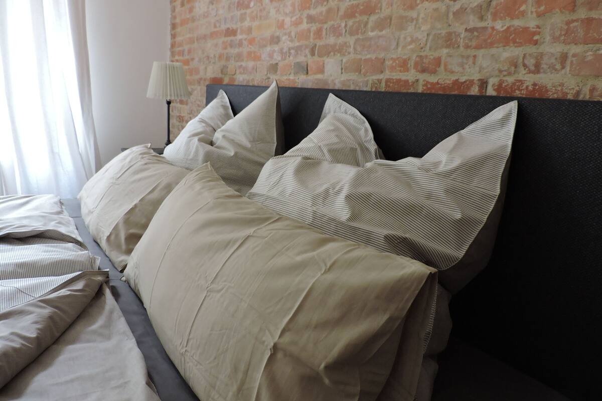 Ein ordentlich gemachtes Bett mit vielen Kissen und einem Kopfteil an einer Ziegelwand.