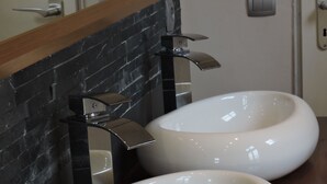 Un baño con dos lavamanos grandes y ovalados, una encimera de madera, un salpicadero de azulejos negros y una toalla adornada con una cinta blanca.