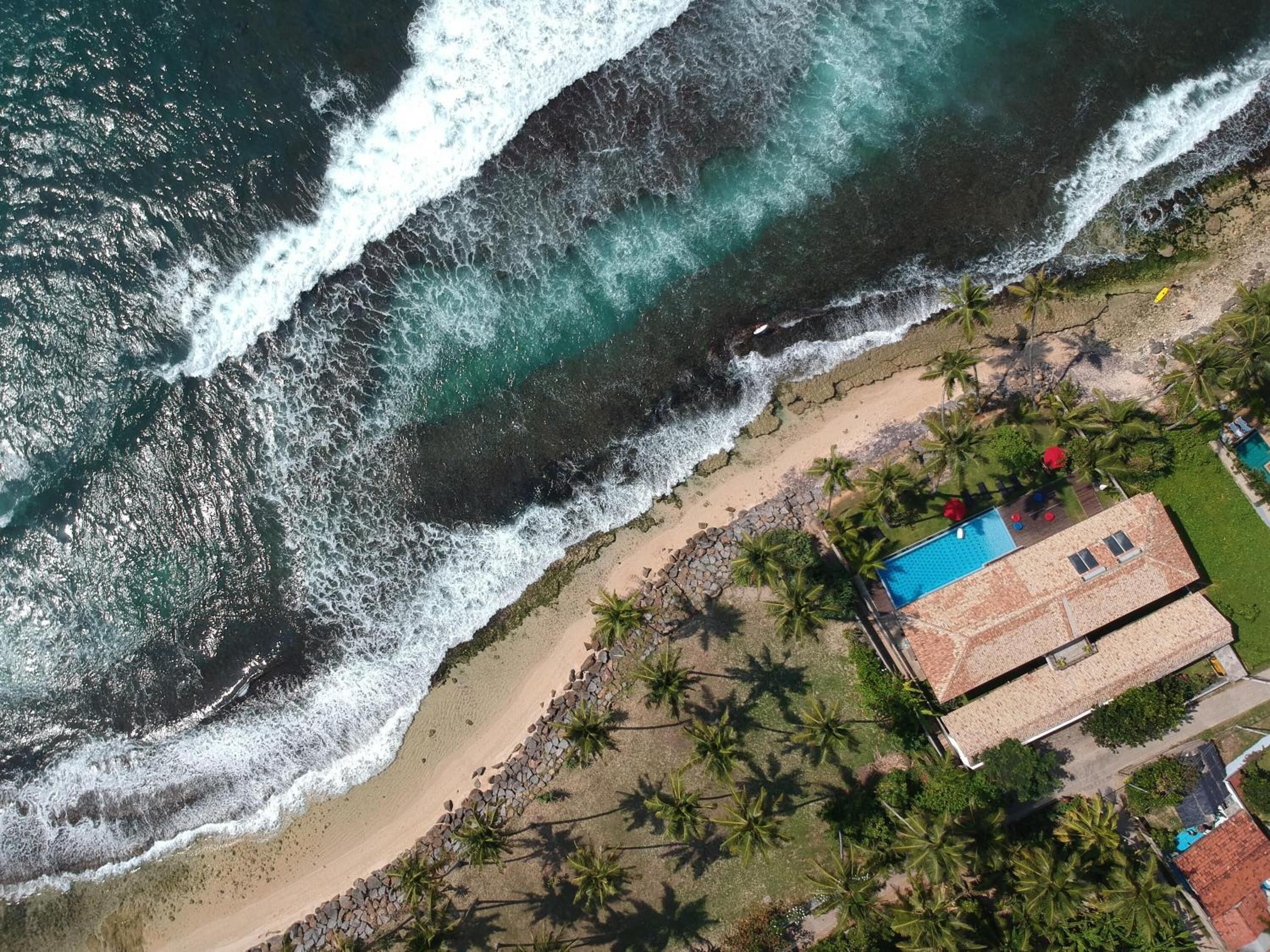 An aerial view of a coastal property with a swimming pool, beach access, and palm trees.