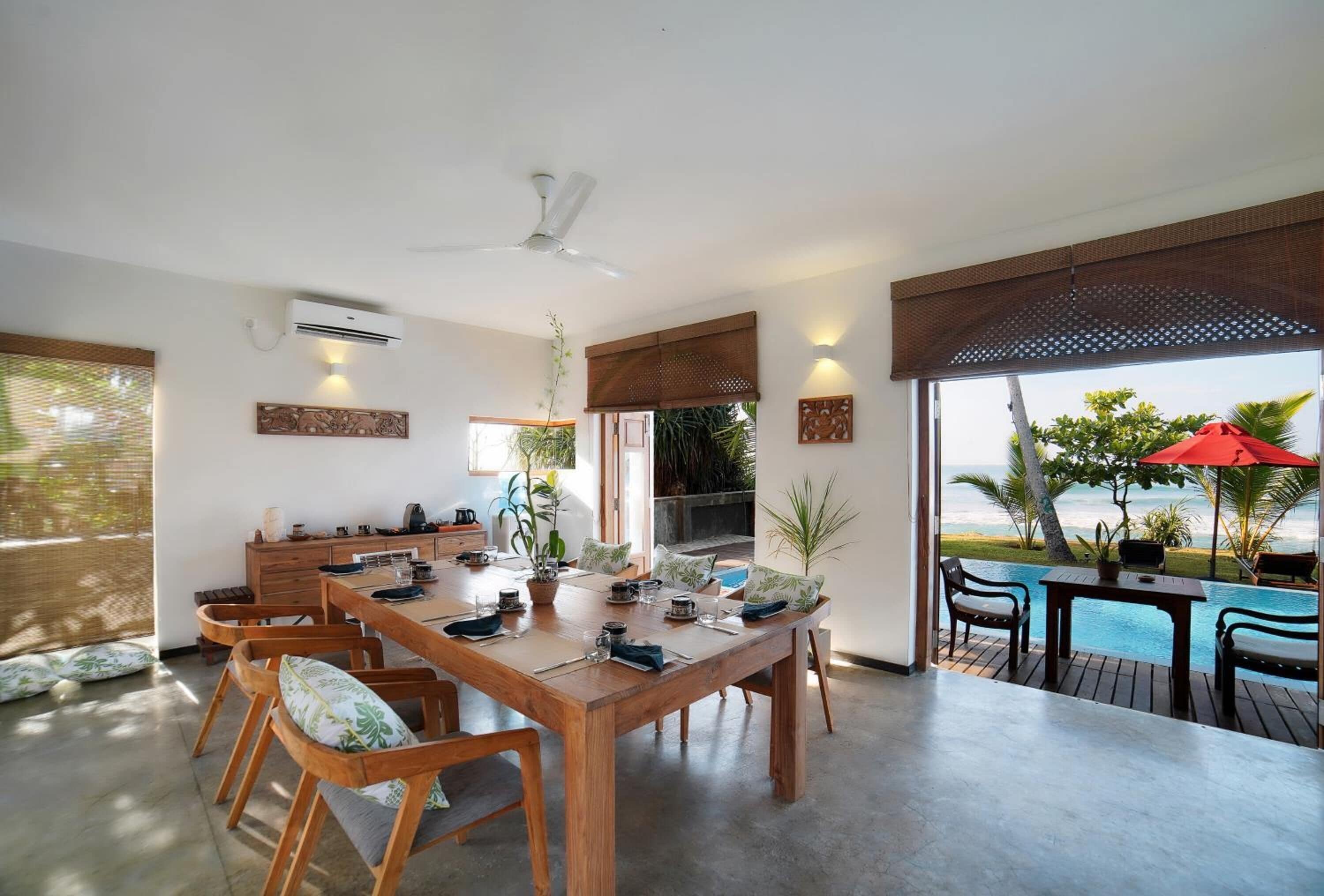 Un amplio comedor con mesa y sillas de madera, ventilador de techo y vista al océano a través de grandes ventanales.