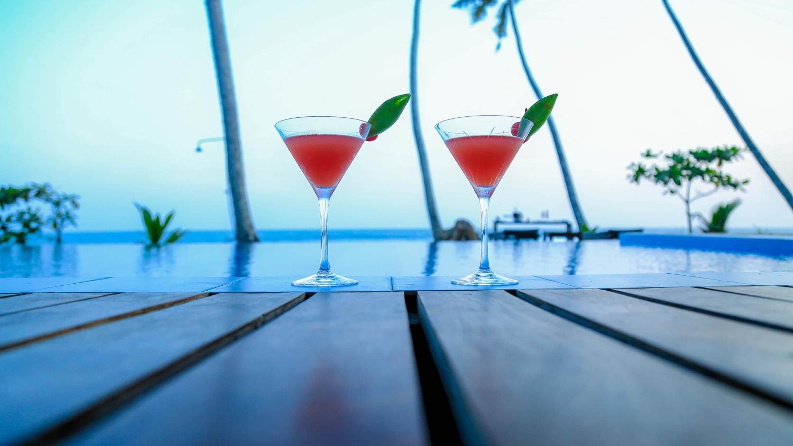 Two martini glasses with a red drink on a wooden deck by a pool with a view of the ocean and palm trees.