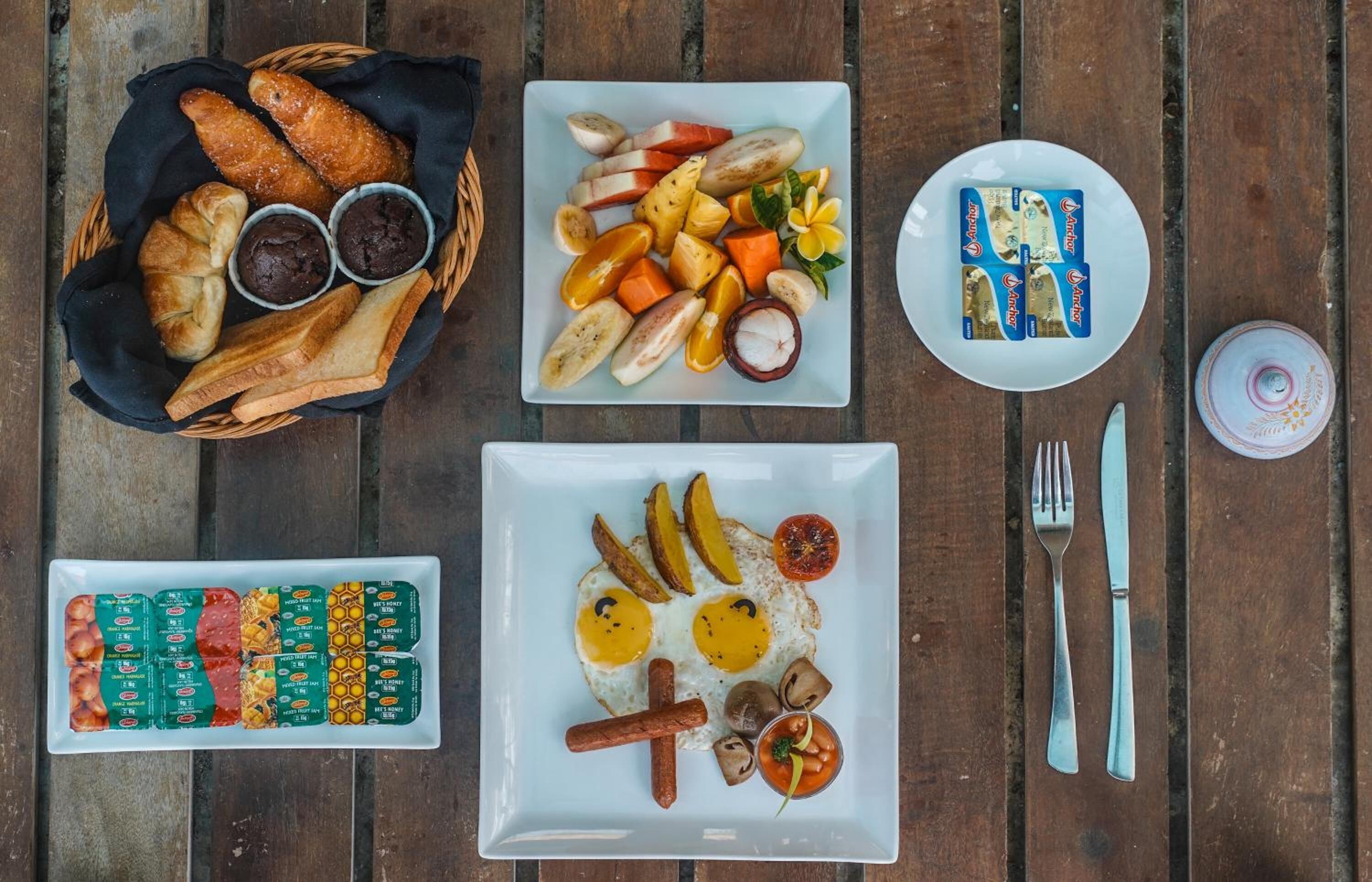 Una mesa de madera con diversos productos para el desayuno, como cruasanes, fruta, huevos, salchichas y condimentos.