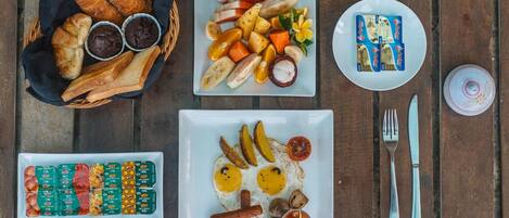 Una mesa de madera con diversos productos para el desayuno, como cruasanes, fruta, huevos, salchichas y condimentos.