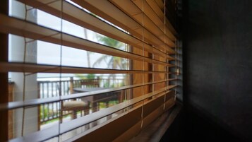 A window with horizontal blinds, a view of an outdoor balcony with a railing, and a glimpse of greenery outside.