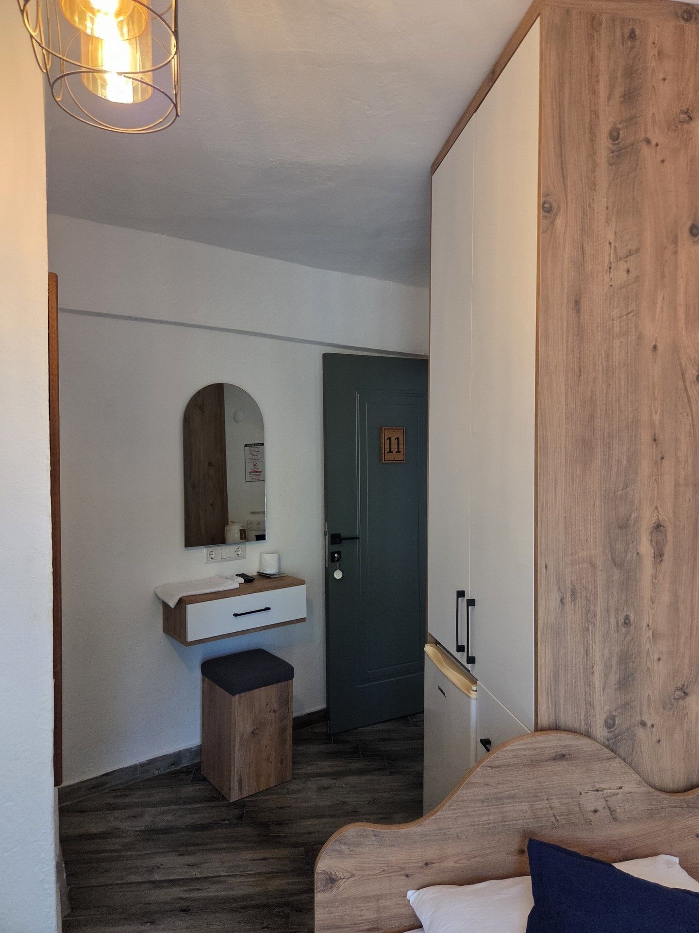 A room with a wooden floor, a bed with a headboard, a bedside table, a mirror, a door with a number, and a wall-mounted light fixture.