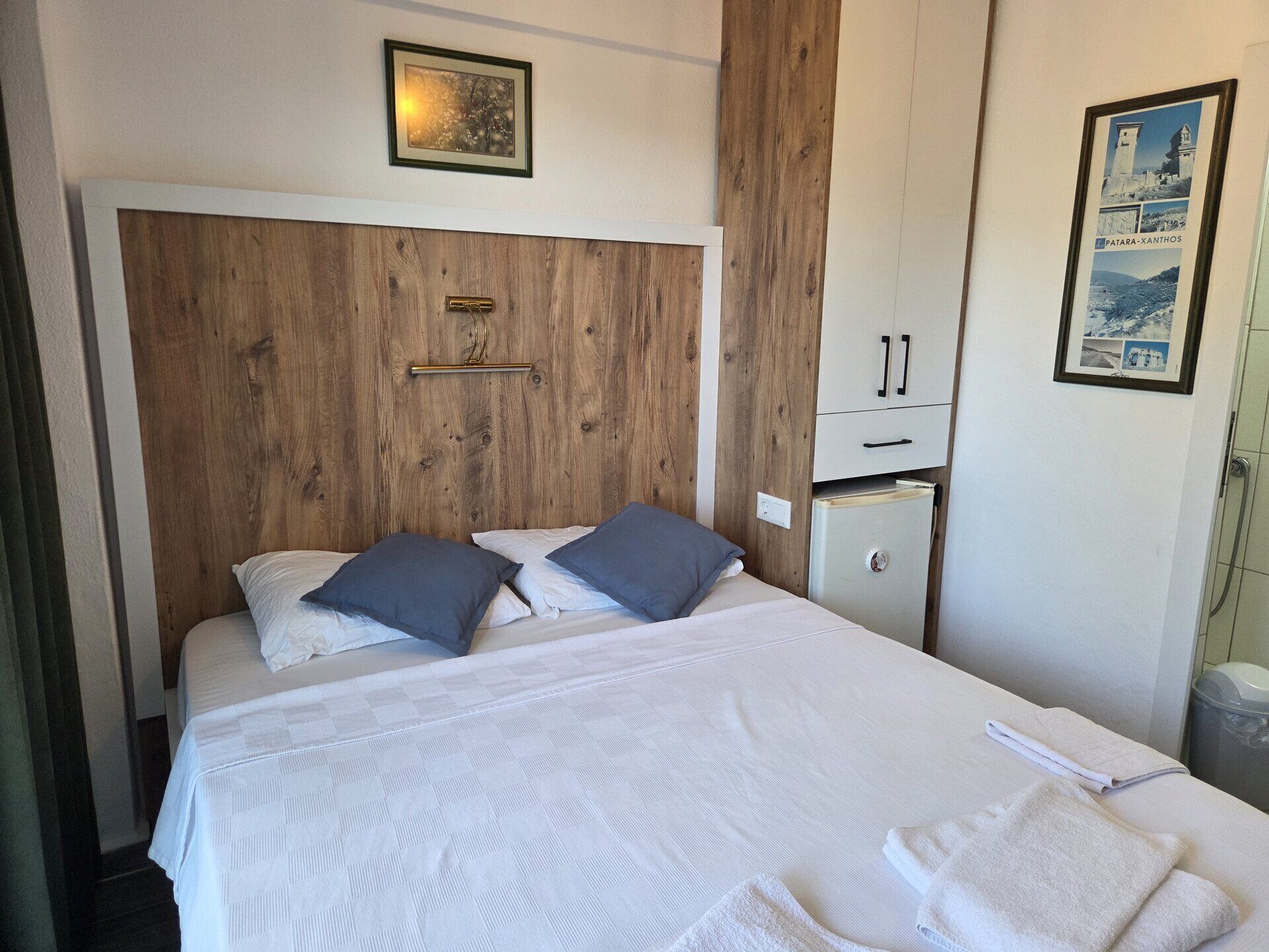 Una habitación de hotel con cabecera de madera, cama con sábanas blancas, dos almohadas, un archivador pequeño y un cartel en la pared.