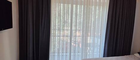 A hotel room with two beds, a TV mounted on the wall, and a view of a balcony through sheer curtains.