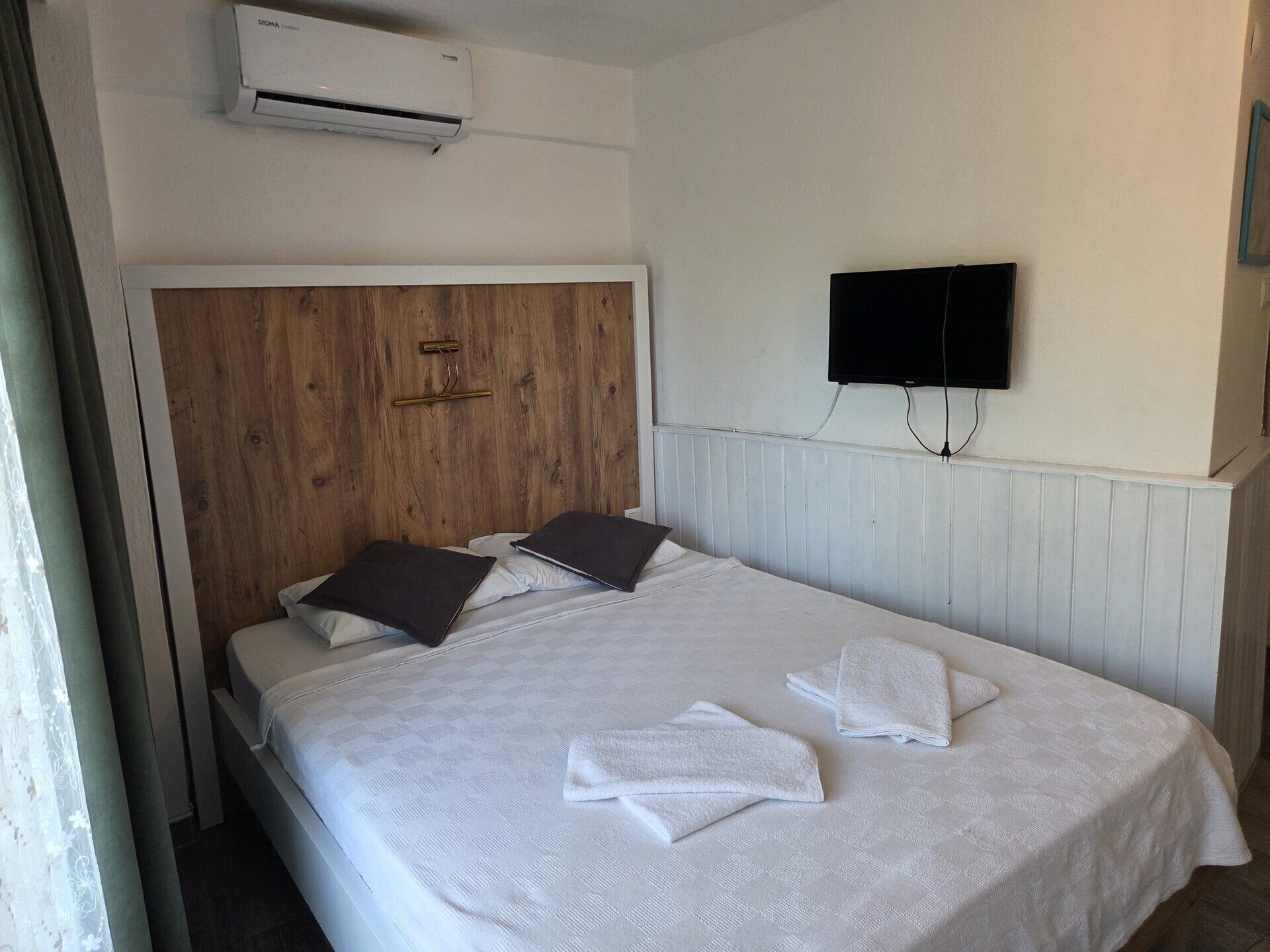 A hotel room with a wooden headboard, a flat-screen TV mounted on the wall, and a bed with white linens and towels.
