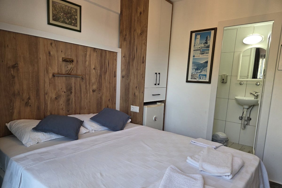 Hotellihuone, jossa on sänky, pieni kaappi, peili ja seinällä oleva kehystetty kuva.
