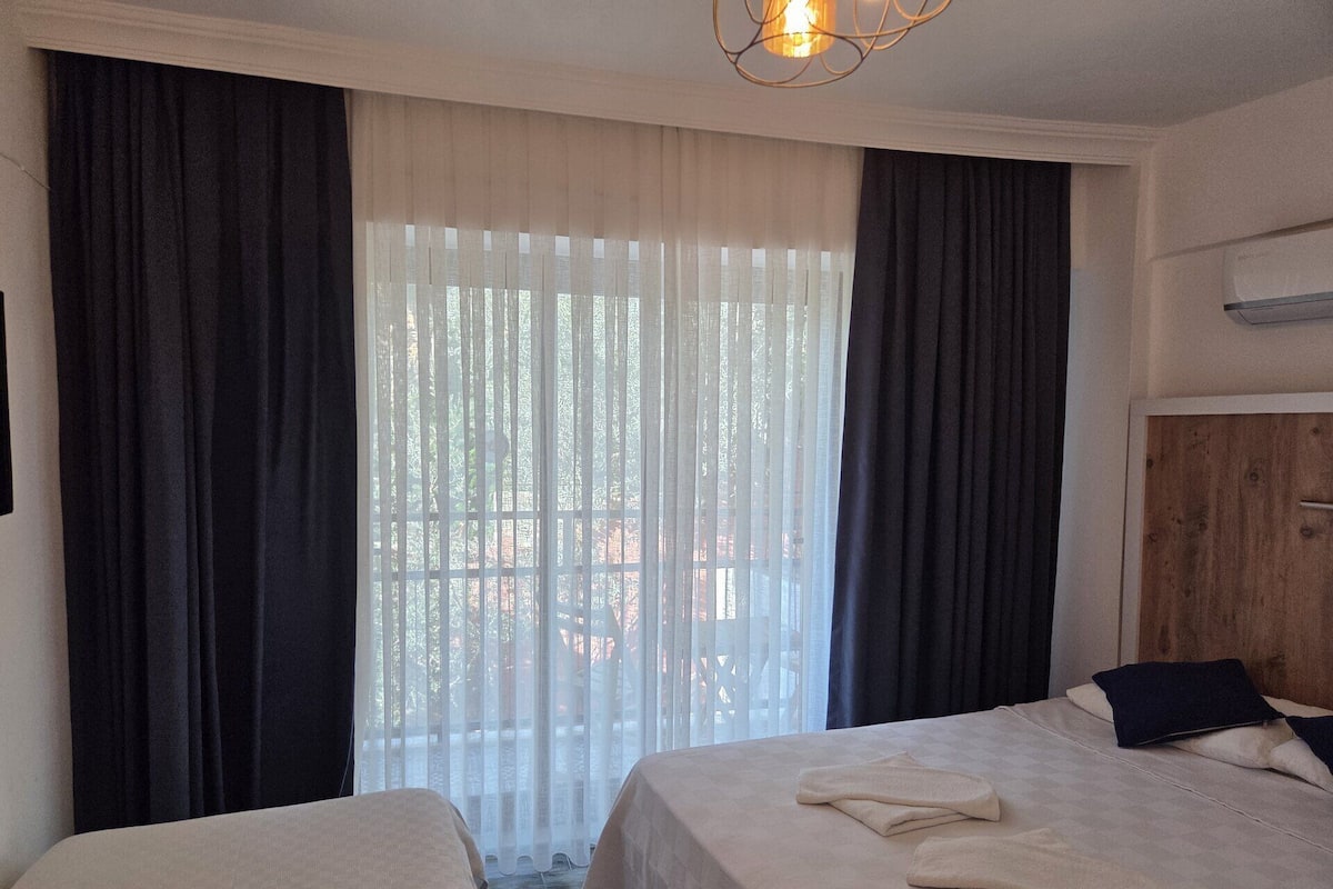Hotellihuone, jossa on kaksi sänkyä, puinen vaatekaappi, ikkuna, jossa on ohuet verhot, ja kattovalaisin.