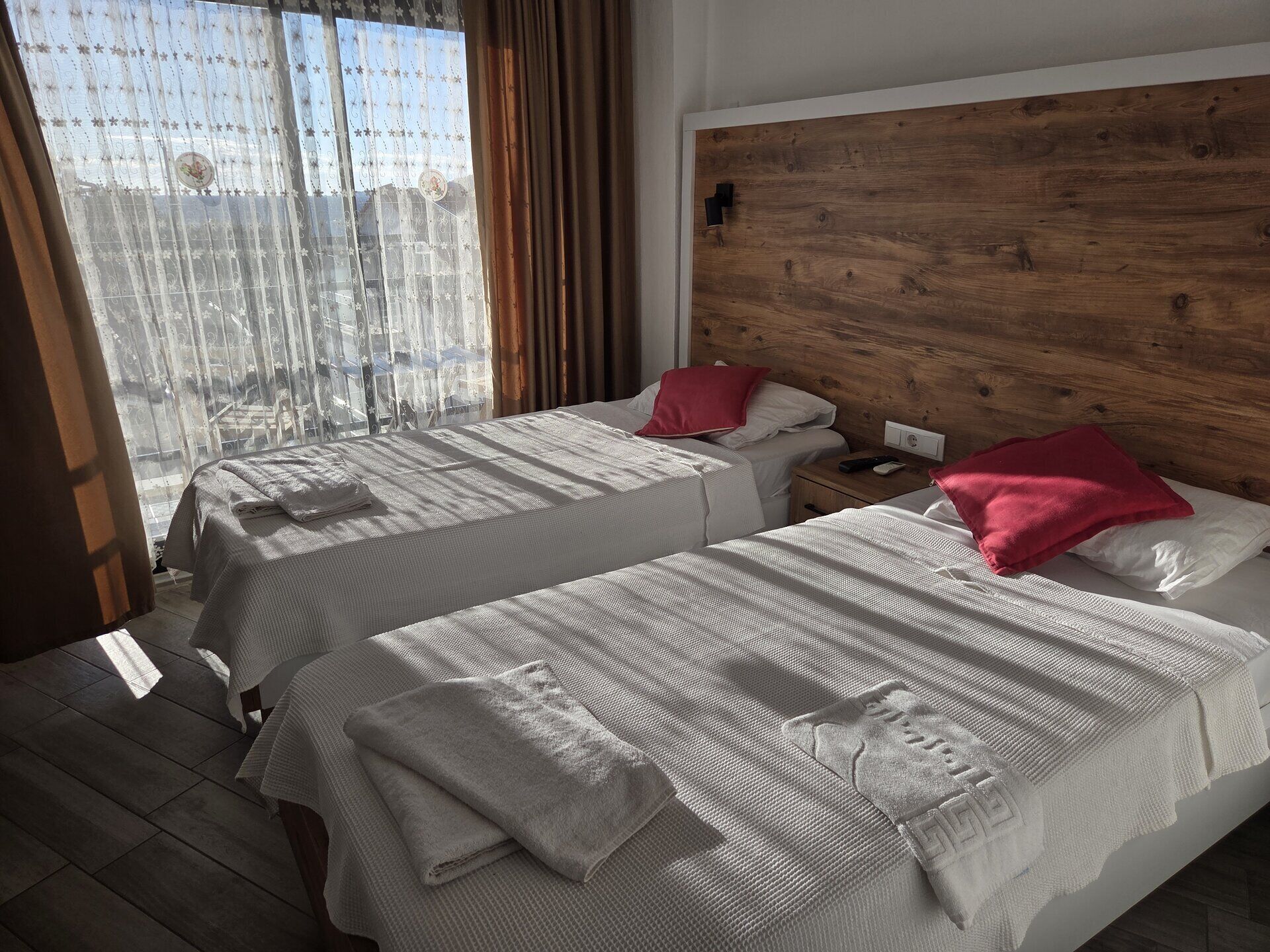 A hotel room with two beds, wooden headboard, white bedding, and a view through sheer curtains.