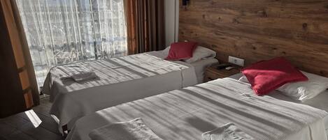 A hotel room with two beds, wooden headboard, white bedding, and a view through sheer curtains.