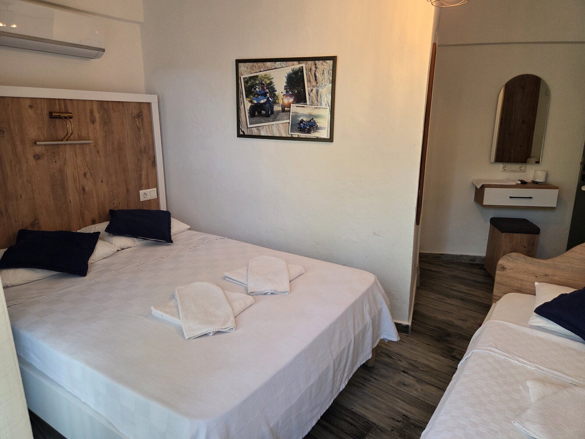 Habitación de hotel con cabecera de madera, cama con sábanas blancas, mesita de noche pequeña, espejo y un cuadro colgado en la pared.