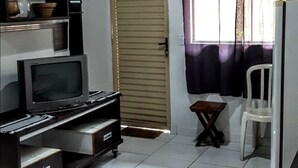 Una habitación con una cama, un televisor sobre un mueble bajo, una mesita, una silla, una puerta y una ventana con cortinas.