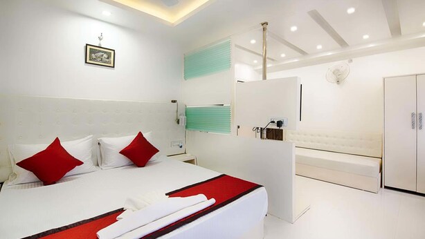 A modern hotel room with a large bed, red pillows, a white headboard, a bedside table, a mirror, a wardrobe, and a bathroom with a bathtub and a toilet. - Hotel Venkatesh Regency (Siliguri)