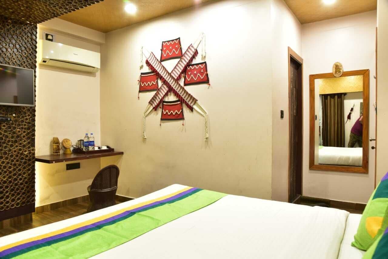 A hotel room with a bed, a desk with a chair, a wall-mounted air conditioner, and a mirror with a curtain.