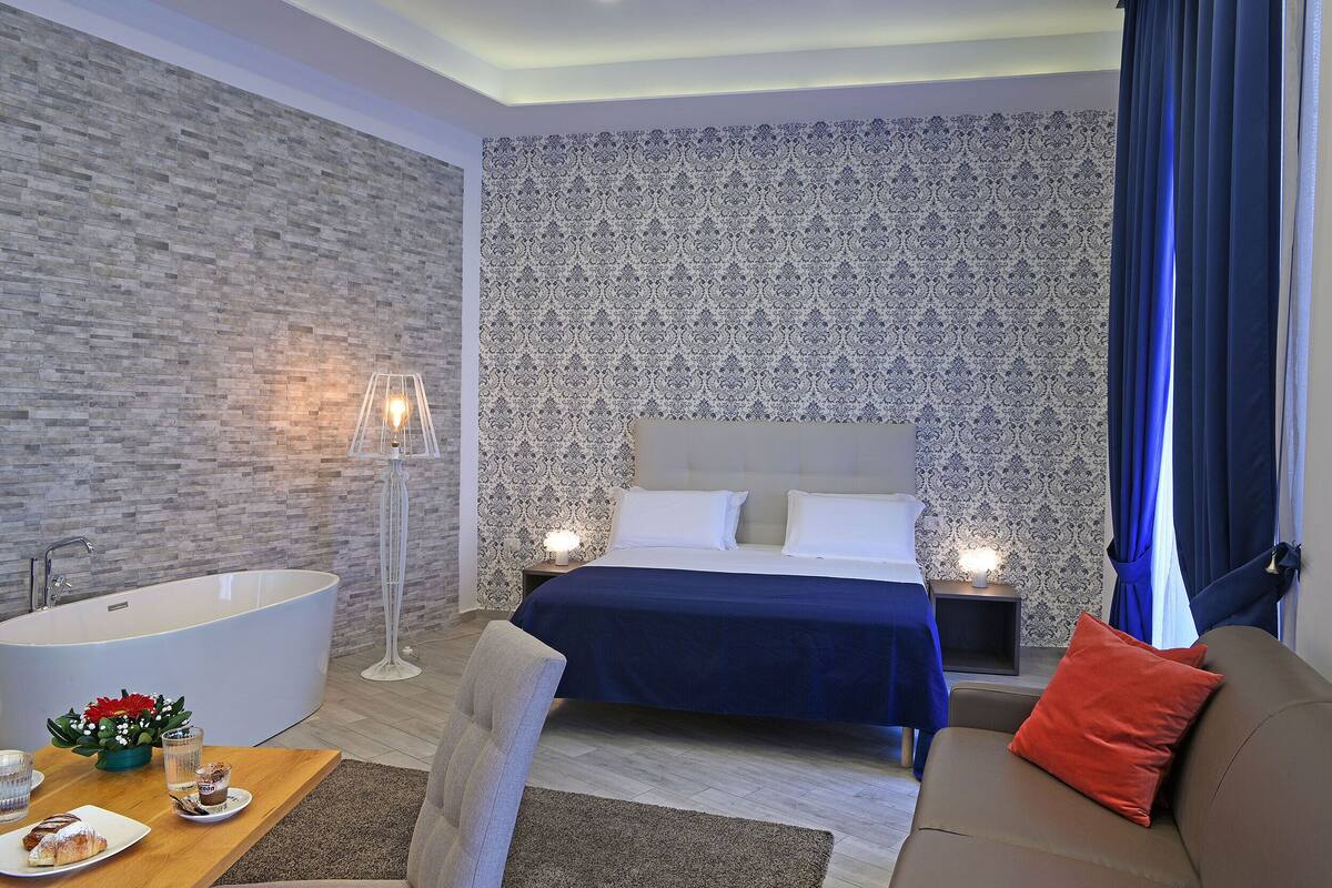 Una habitación de hotel moderna con una bañera grande, una cama con cubrecama azul oscuro, un sofá con un cojín rojo y una pequeña mesa con un plato de pasteles y un vaso de agua.