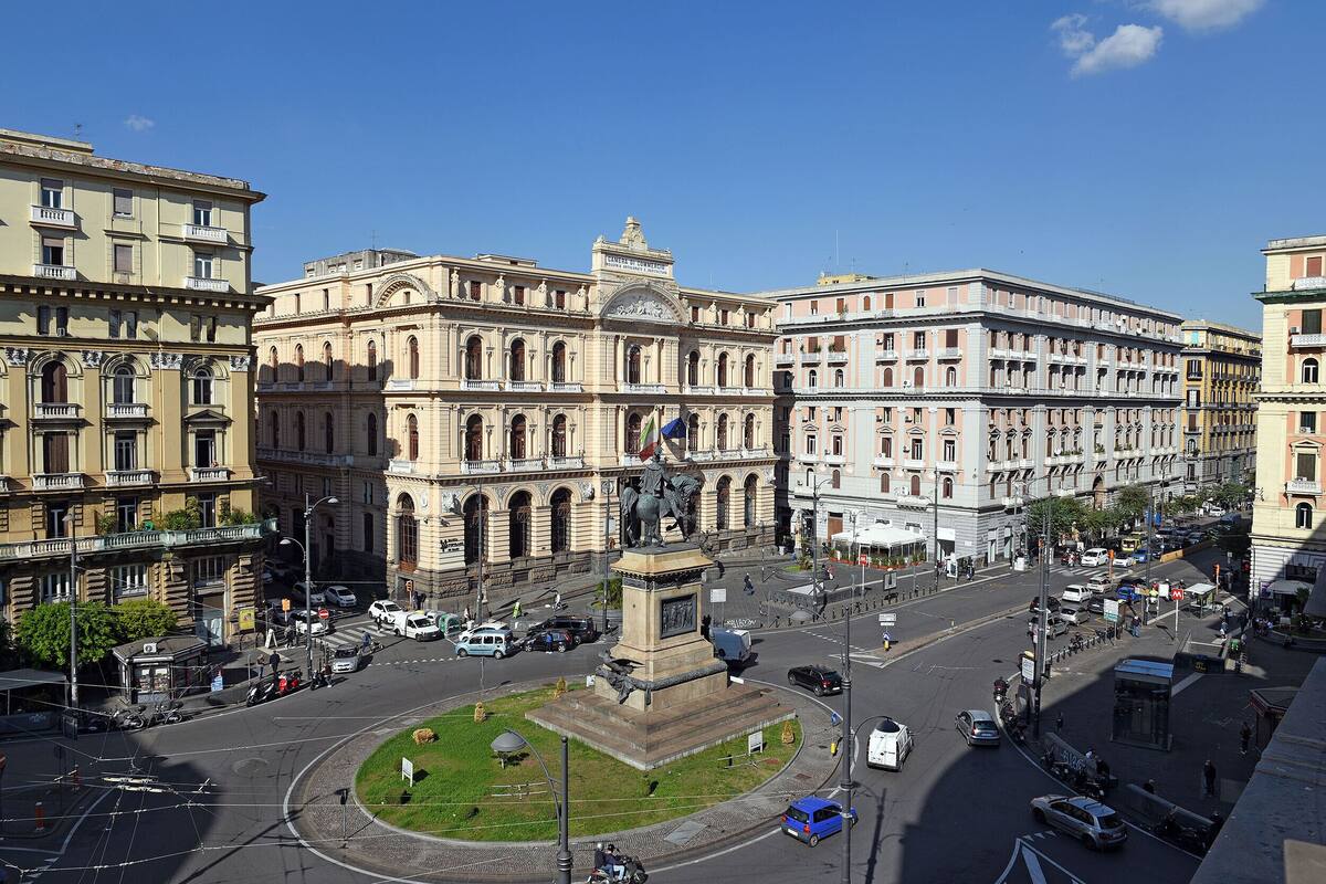 Un edificio histórico con arquitectura clásica, un monumento central y una calle urbana concurrida.