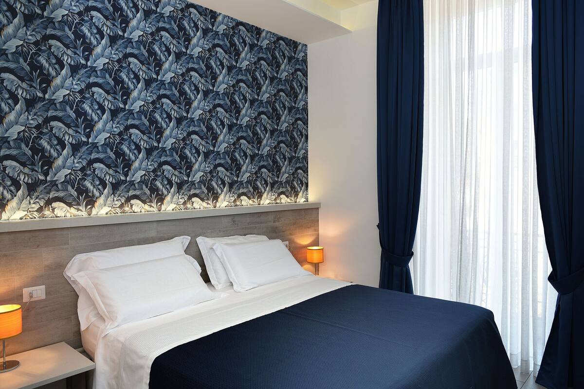 Habitación de hotel con una cama, ropa de cama blanca, una mesita de noche con lámpara y una pared con estampado de hojas.