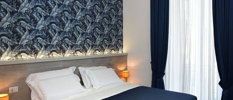 A hotel room with a bed, white linens, a nightstand with a lamp, and a wall with a leaf pattern.