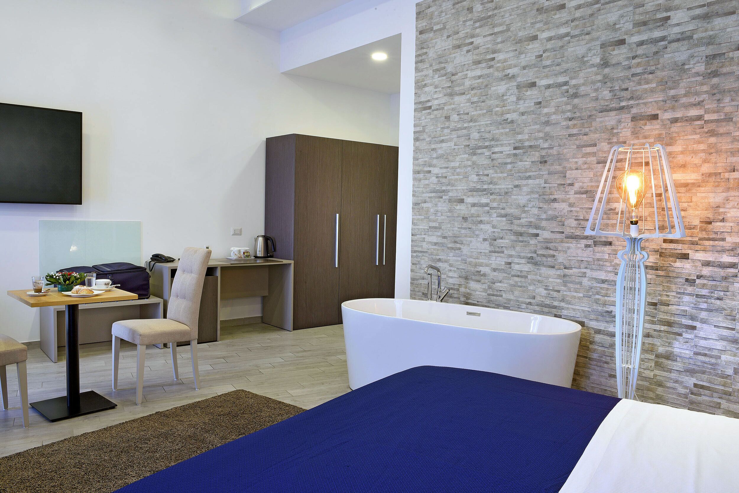 A modern hotel room with a large bed, a desk with a chair, a small table with a plant, a wardrobe, and a bathtub.