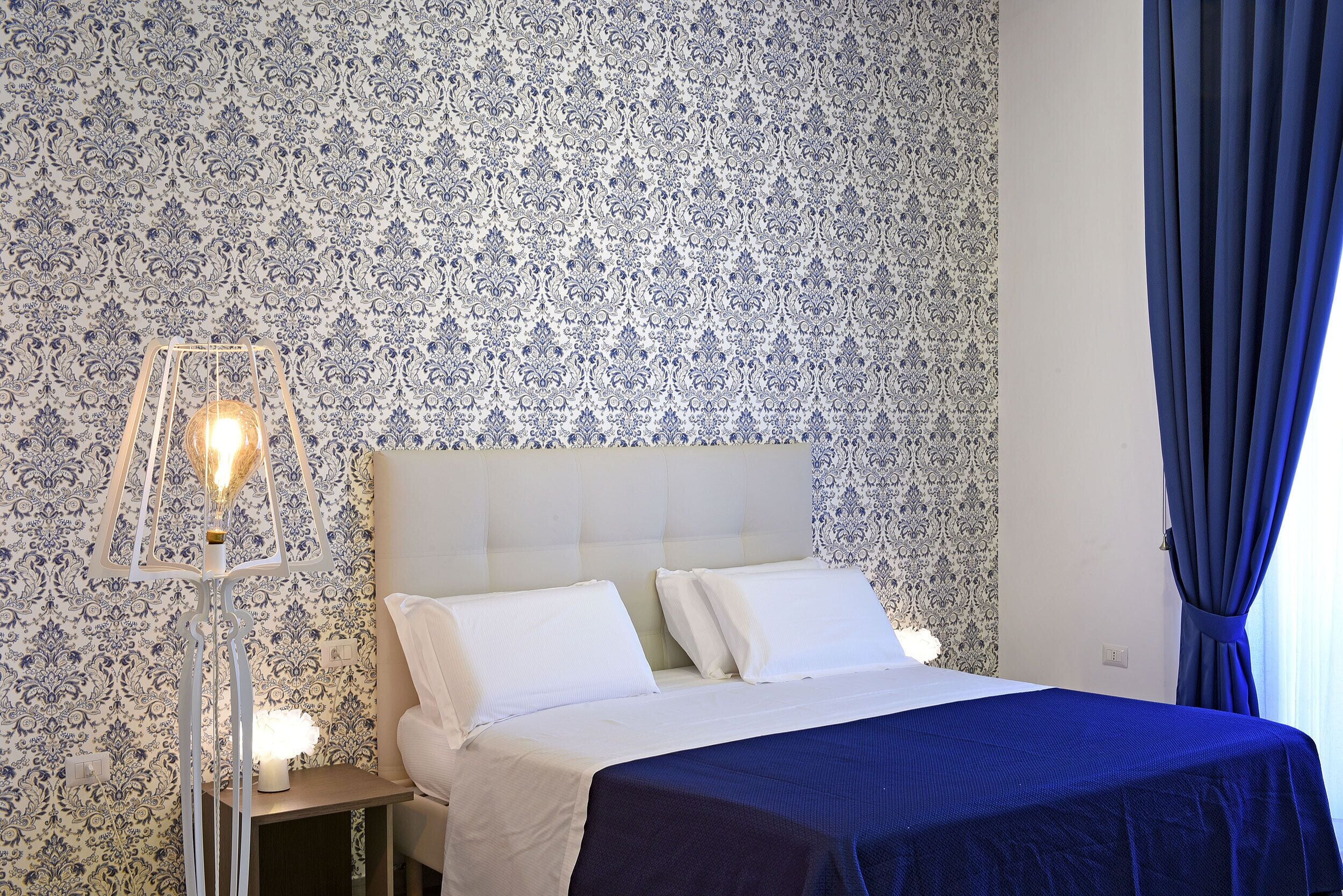 Una cama bien hecha con sábanas blancas y una colcha azul, una mesita de noche con una lámpara y un papel tapiz estampado.