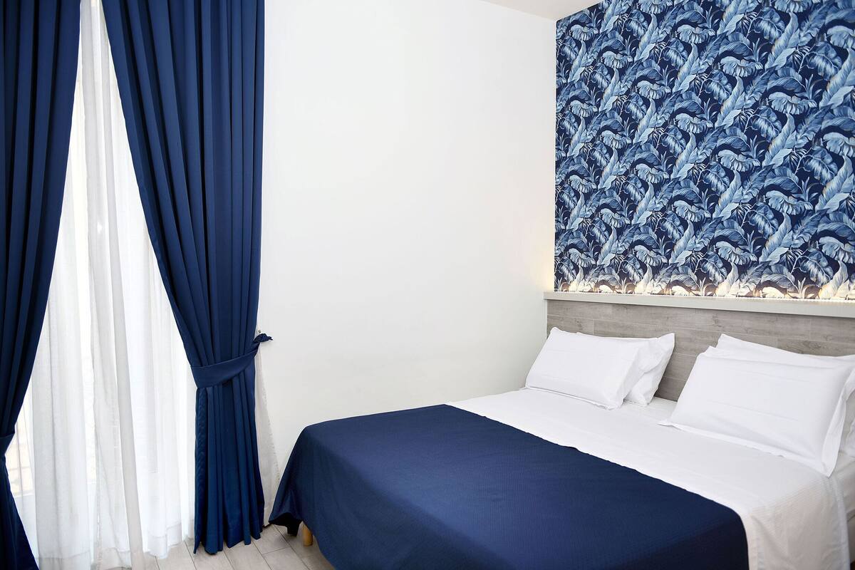 Habitación de hotel con cama, cortinas blancas y pared con diseño azul.