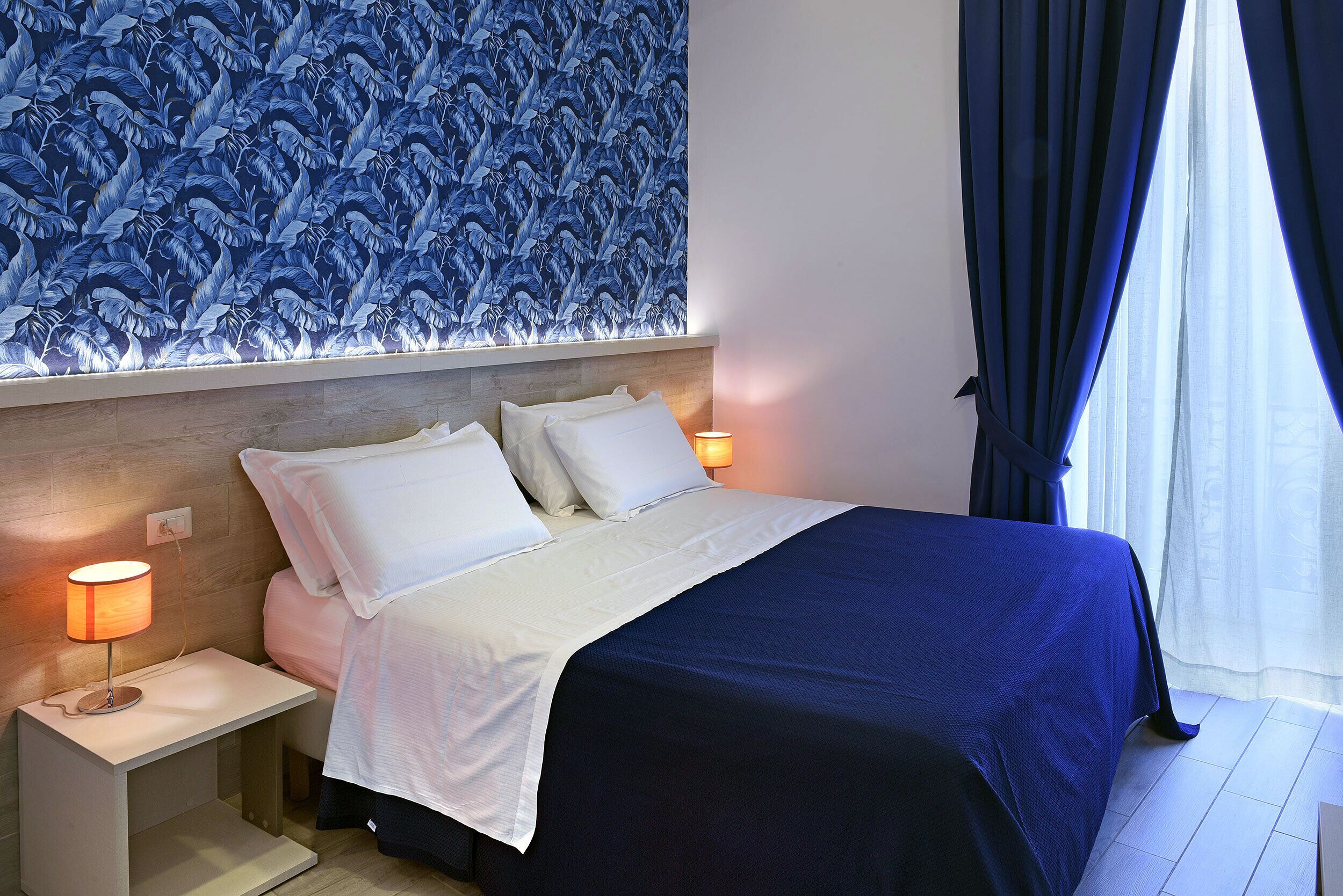Una cama bien hecha con ropa de cama blanca y azul, una mesita de noche con una lámpara y una pared con un estampado de hojas azules.