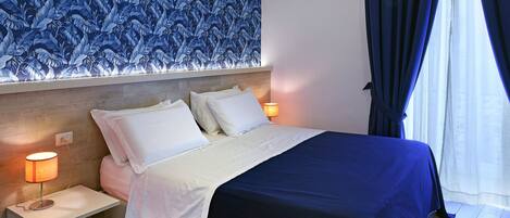 Una cama bien hecha con ropa de cama blanca y azul, una mesita de noche con una lámpara y una pared con un estampado de hojas azules.