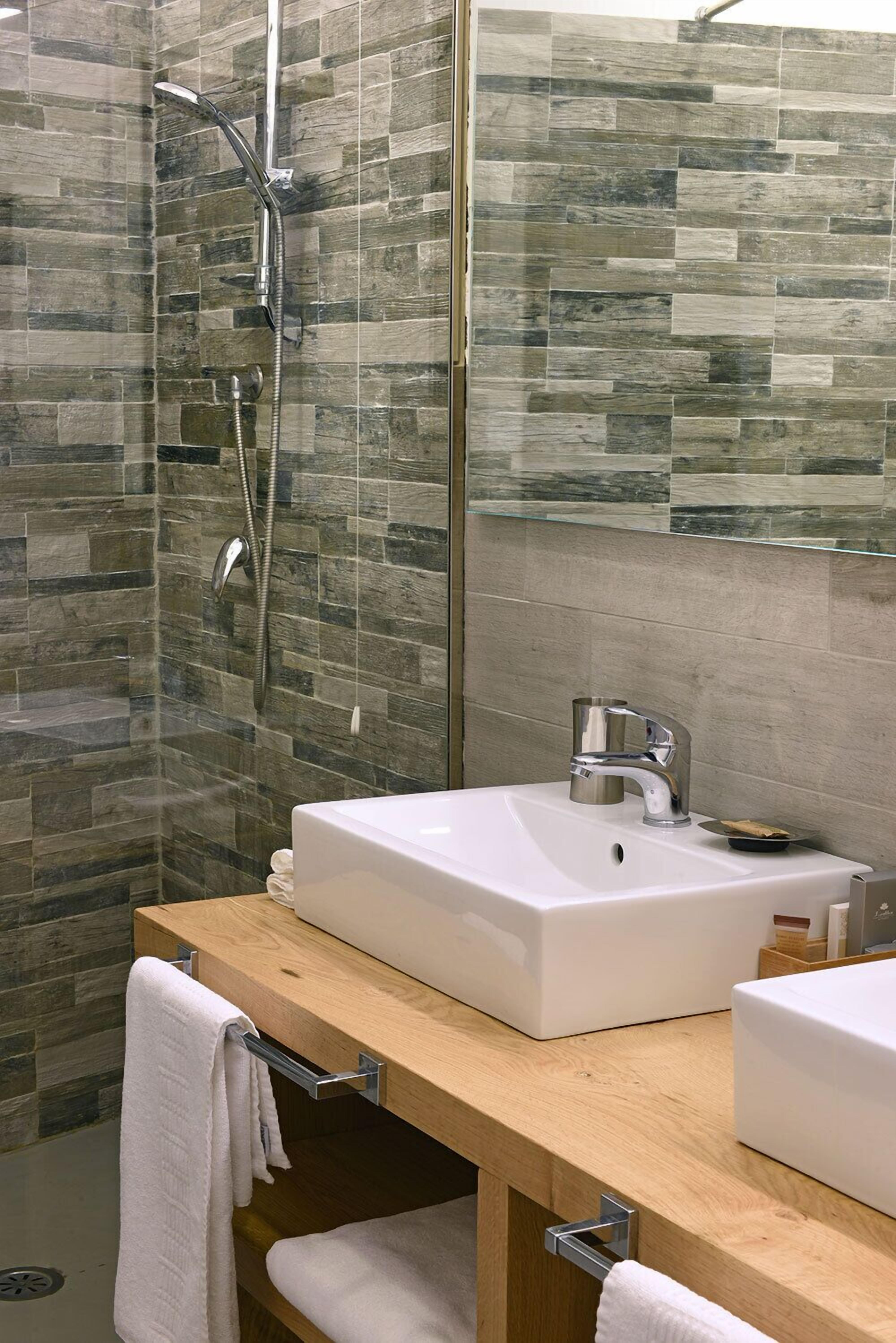 Een moderne badkamer met een stenen muur, een douchecabine van glas, een witte wastafel en een houten wastafelmeubel.