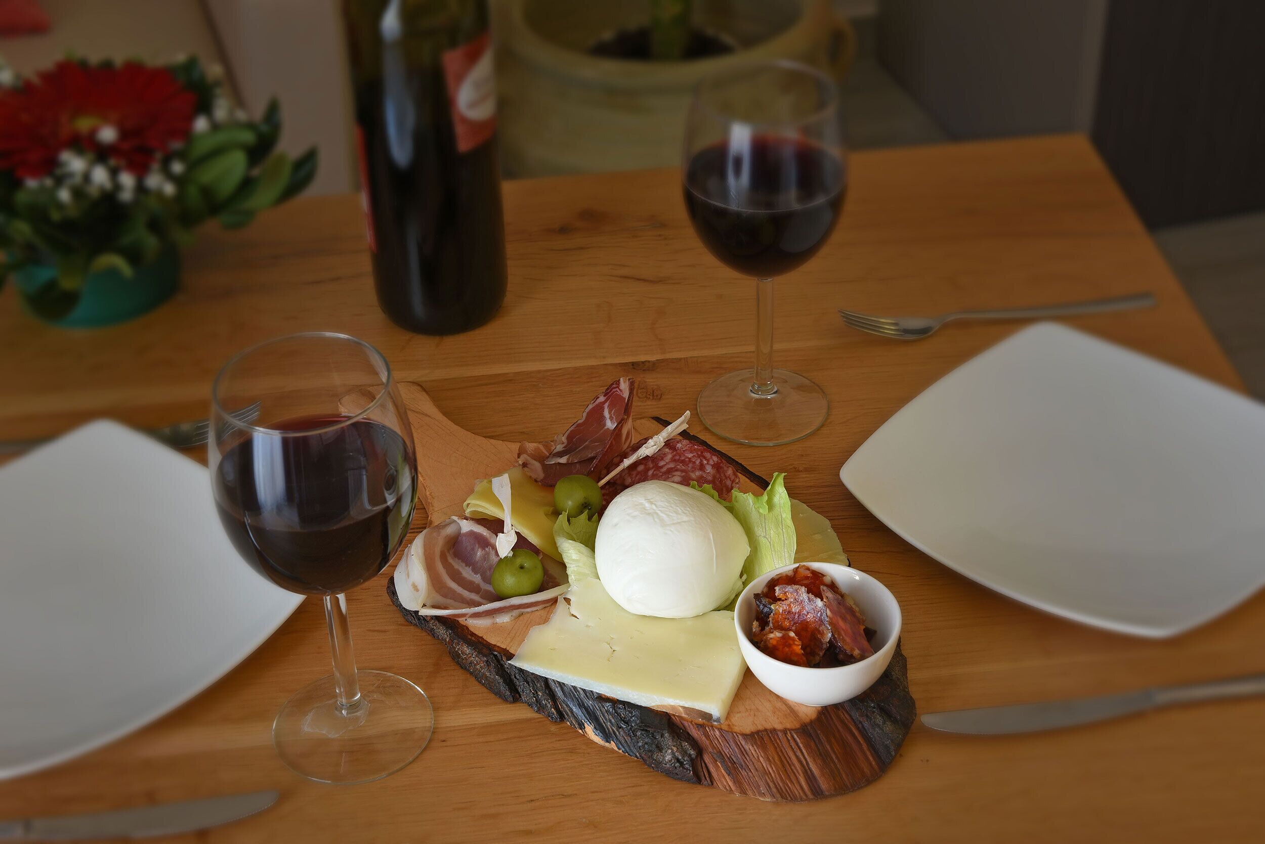 A wooden table with a cheese board, two glasses of red wine, and a bottle of wine in the background.