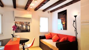 Un espacio habitable compacto con un sofá, una mesa de comedor con sillas, una lámpara de pie y un aire acondicionado de pared.