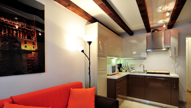 Cocina moderna con microondas, pileta y armarios de acero inoxidable. En la zona de estar, hay un sofá rojo con cojines naranja.