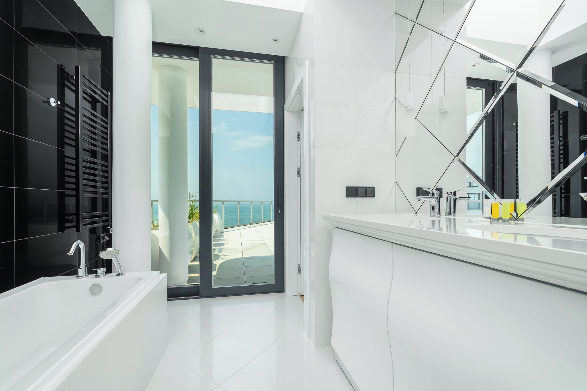 A modern bathroom with a large bathtub, a glass door leading to a balcony, and a large mirror.