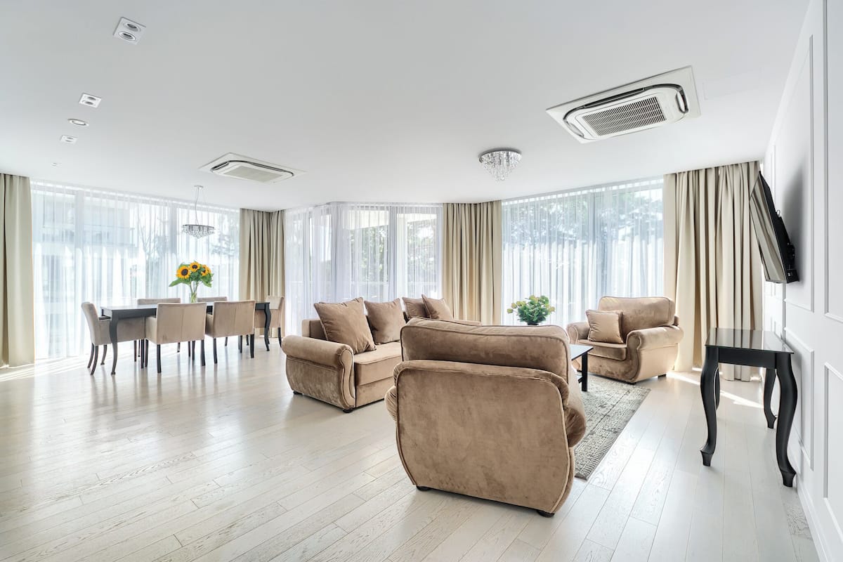 Ein großzügiger Wohnraum mit Essbereich, beigen Sofas, einem schwarzen Tisch und großen Fenstern mit Vorhängen.